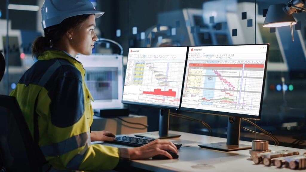 A planner in a hard hat and high-visibility jacket reviewing Powerproject Gantt charts on two monitors