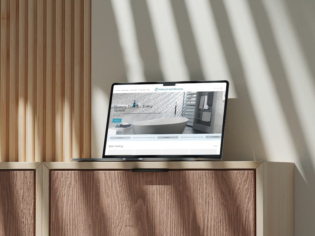 A laptop on a wooden cabinet showing the Forazzi Bathrooms homepage, with a freestanding bath