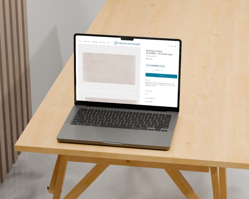 A laptop on a light wood table showing the Bottega Caliza porcelain tile product page