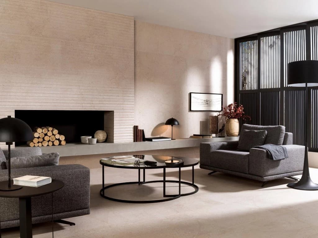 A living room finished in pale stone tiles, with a ribbed fireplace, grey seating and black lighting